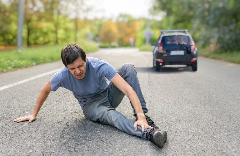 Man in gray clothing sitting on a road, holding his injured ankle, with a black car in the background.