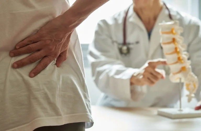 Patient holding lower back in pain with doctor explaining spine model in background.