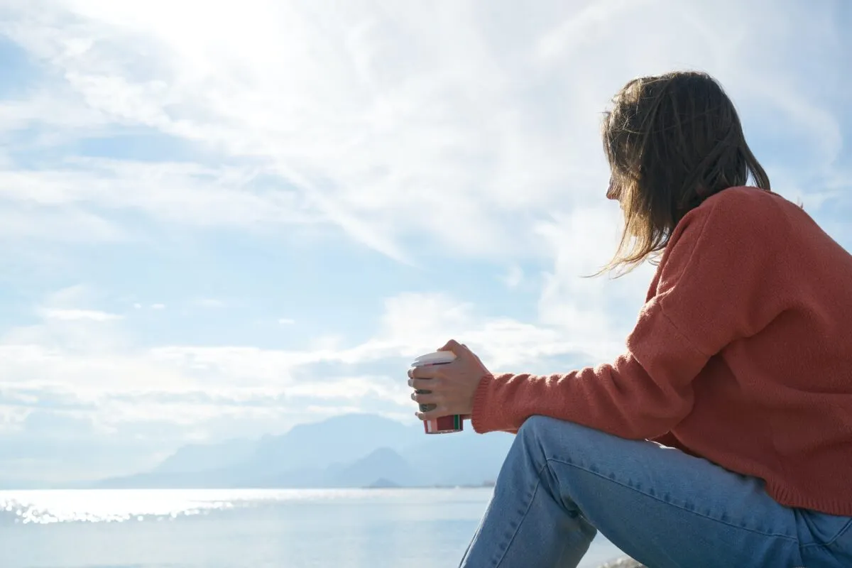 Woman in red sweater and jeans holding a coffee cup, sitting by a calm sea under a cloudy sky.