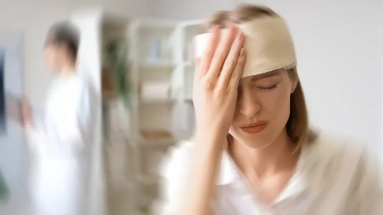 Woman with a head bandage holding her forehead in pain, blurred medical professional in background.
