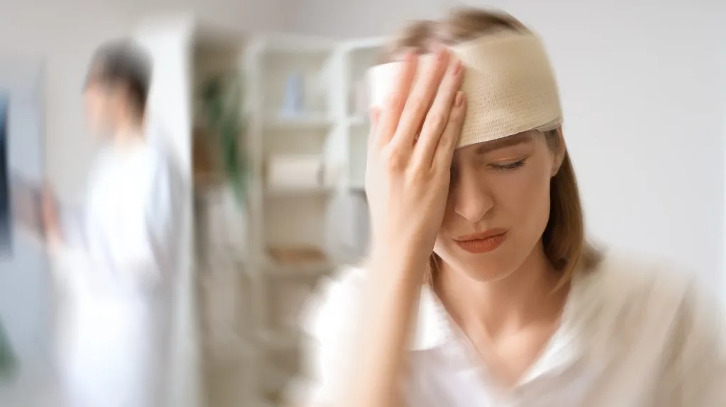 Woman with a head bandage holding her forehead in pain, blurred medical professional in background.